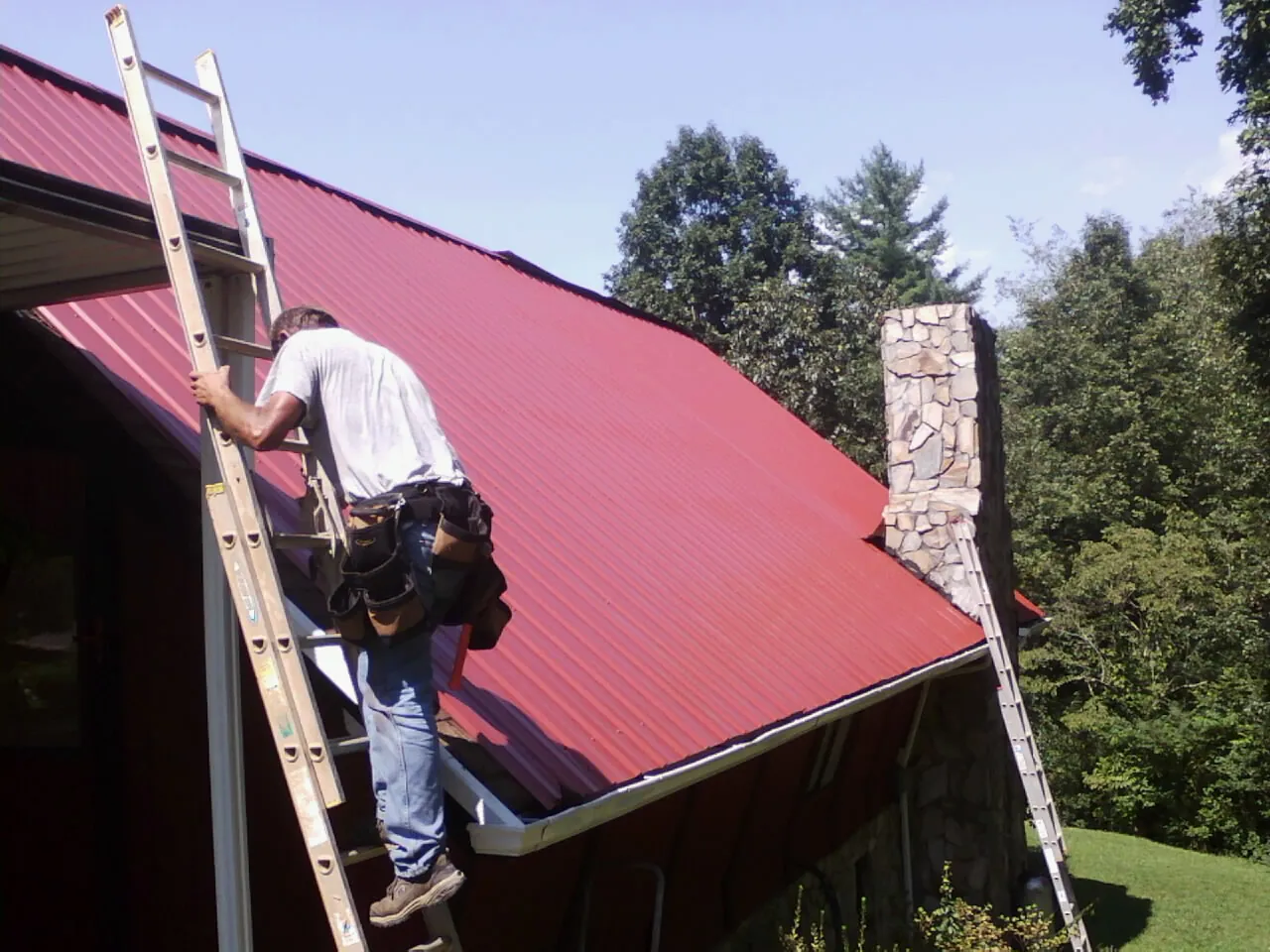 Roofer on a ladder beside a red metal roof and stone chimney — Soffit & Fascia Repair by Roofing Brothers in Rahway